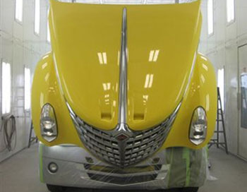 Front-facing yellow truck inside a body repair shop with white walls and two rows of windows.